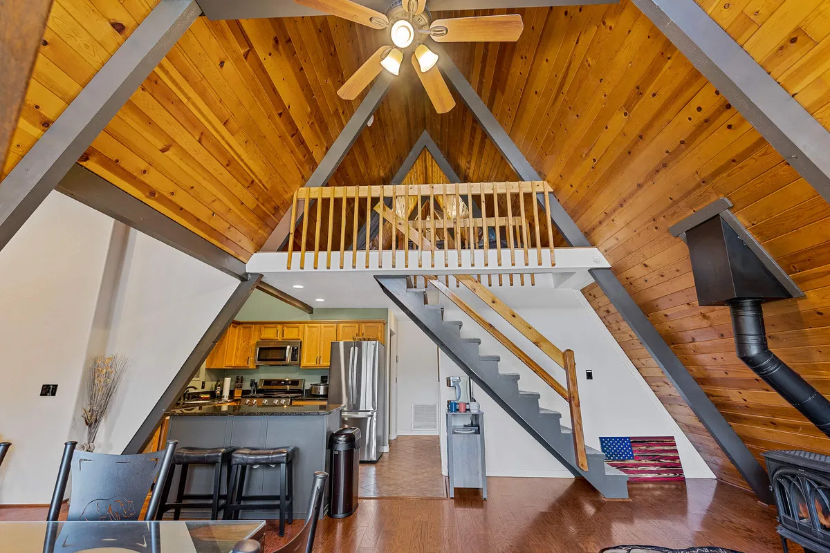 A cozy interior view with a vaulted wooden ceiling, a staircase leading to a loft, and a kitchen and dining area below. The loft area is visible, suggesting a perfect spot for relaxing or gaming above the main living space.