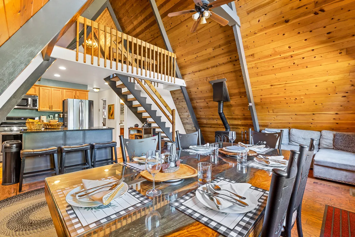 An inviting open-plan interior view with a large dining table set for a meal, a staircase leading to a loft, and a glimpse of the kitchen area. The setting suggests warmth and togetherness, perfect for shared meals and group activities.