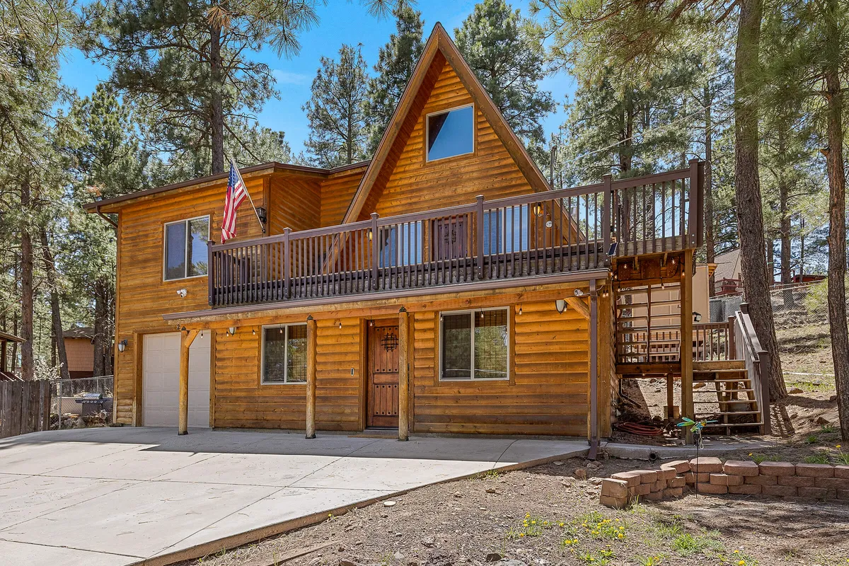 This exterior shot captures the wooden cabin surrounded by tall pine trees, with a large balcony deck adorned with an American flag. The setting exudes a harmonious blend of rustic architecture and natural woodland scenery, hinting at opportunities for morning coffee or post-adventure relaxation outdoors.