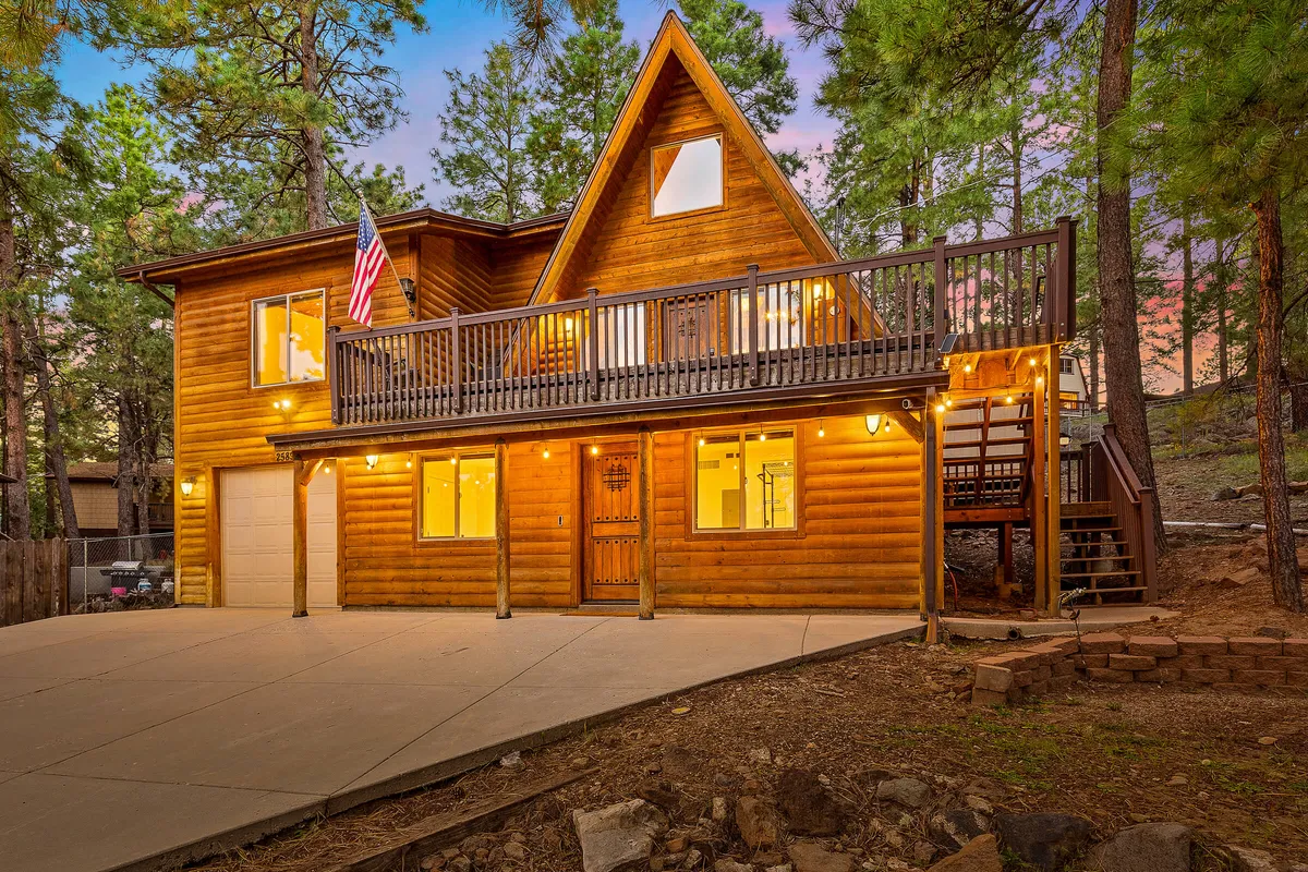 An inviting exterior view of the cabin features a wooden deck with ample seating, large windows, and an American flag. The deck is designed for gatherings and relaxation, situated to take in the natural surroundings and sunsets, signaling an ideal spot for socializing or unwinding after a day of adventure.