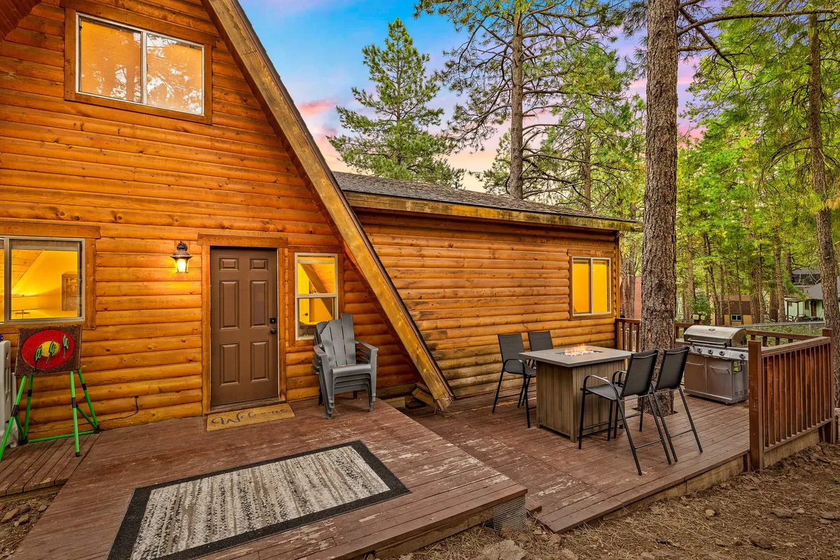 A view of the cabin's outdoor dining table set on a wooden deck, with a barbecue grill nearby. The setting is surrounded by nature and ideal for group meals or evening barbecues, blending the comforts of home with the forest environment.