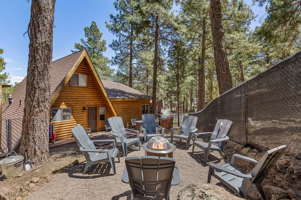 The image features a classic wooden A-frame cabin nestled among tall pine trees, with a welcoming fire pit surrounded by Adirondack chairs in the foreground. The setting evokes warmth and togetherness, perfect for group gatherings beneath the starry Flagstaff sky.