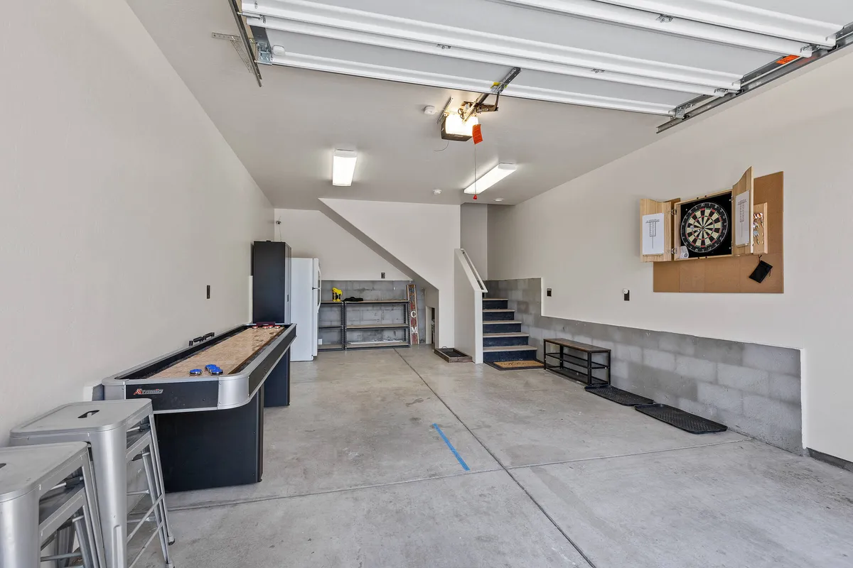 This image centers on a spacious game room featuring a pool table and dartboard, with a visible staircase and a refrigerator tucked to the side. The setting is casual, with warm wood tones and plenty of room for group activities and social gatherings.