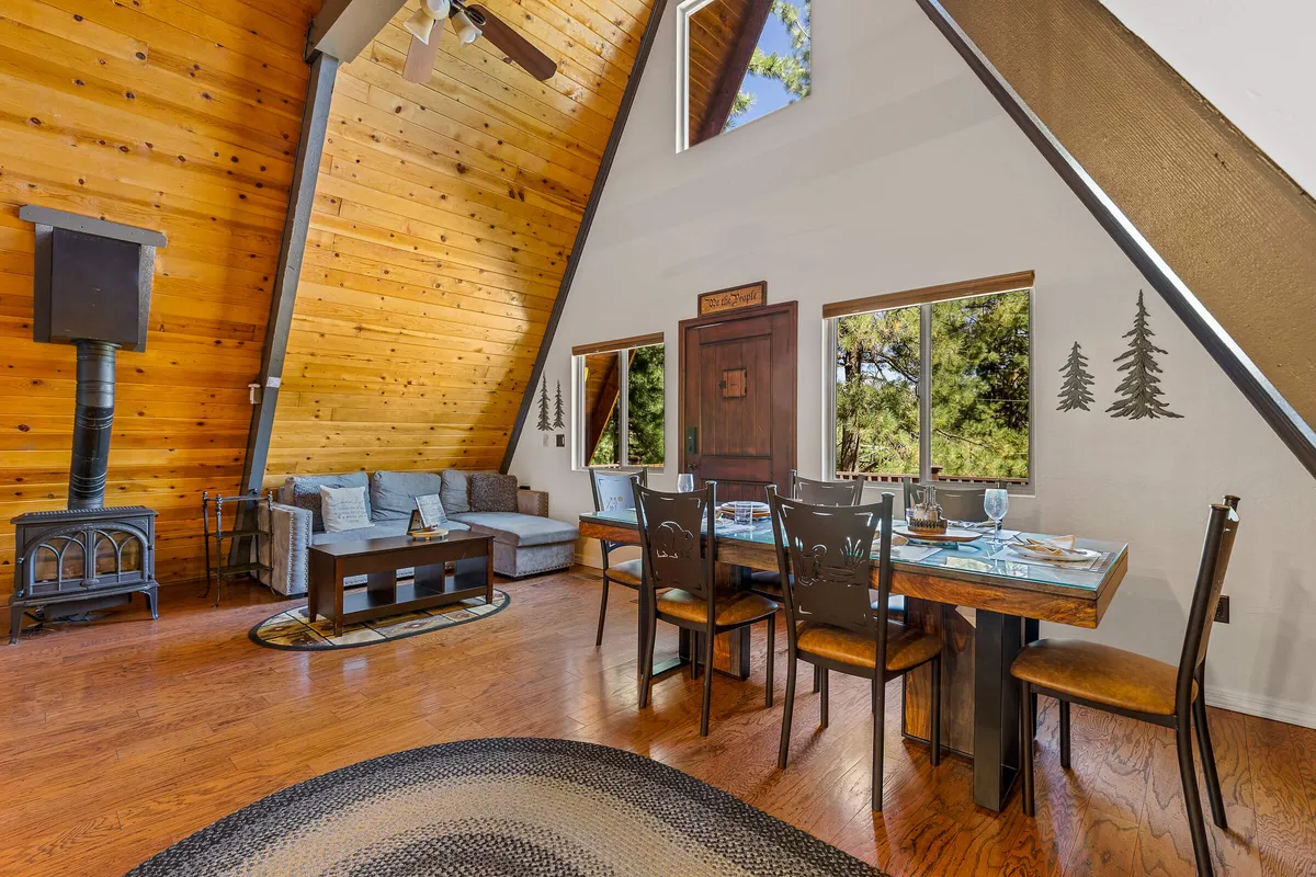 A spacious and inviting living/dining area with an A-frame ceiling, wood-burning stove, large windows offering views of the outdoors, a wooden dining table, and comfortable seating. The atmosphere is warm, blending rustic charm with ample natural light and modern comfort.