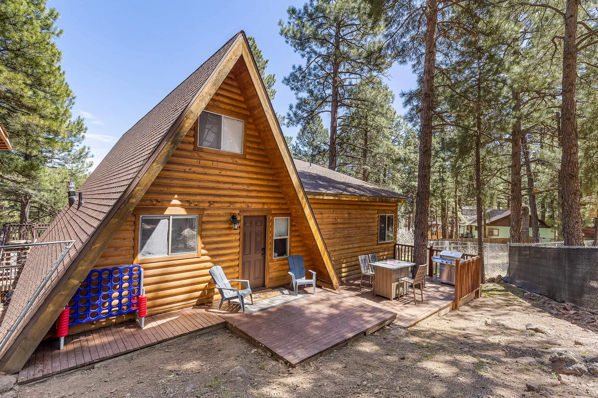 This image features the A-frame structure of the cabin prominently nestled among tall pine trees, with an inviting wooden deck and comfortable outdoor seating area. The scene emphasizes the natural surroundings and serves as a visual gateway to outdoor adventure. The deck's vantage point hints at stargazing, morning coffee, and barbecues, reinforcing connections to the surrounding wilderness.