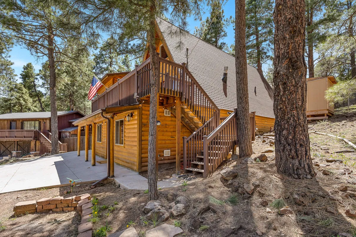 The exterior of a log cabin is framed by a wooden staircase, American flag, and lush greenery, inviting families to step outside for relaxation or embark on nature walks. The deck and open yard suggest space for games and barbecues.
