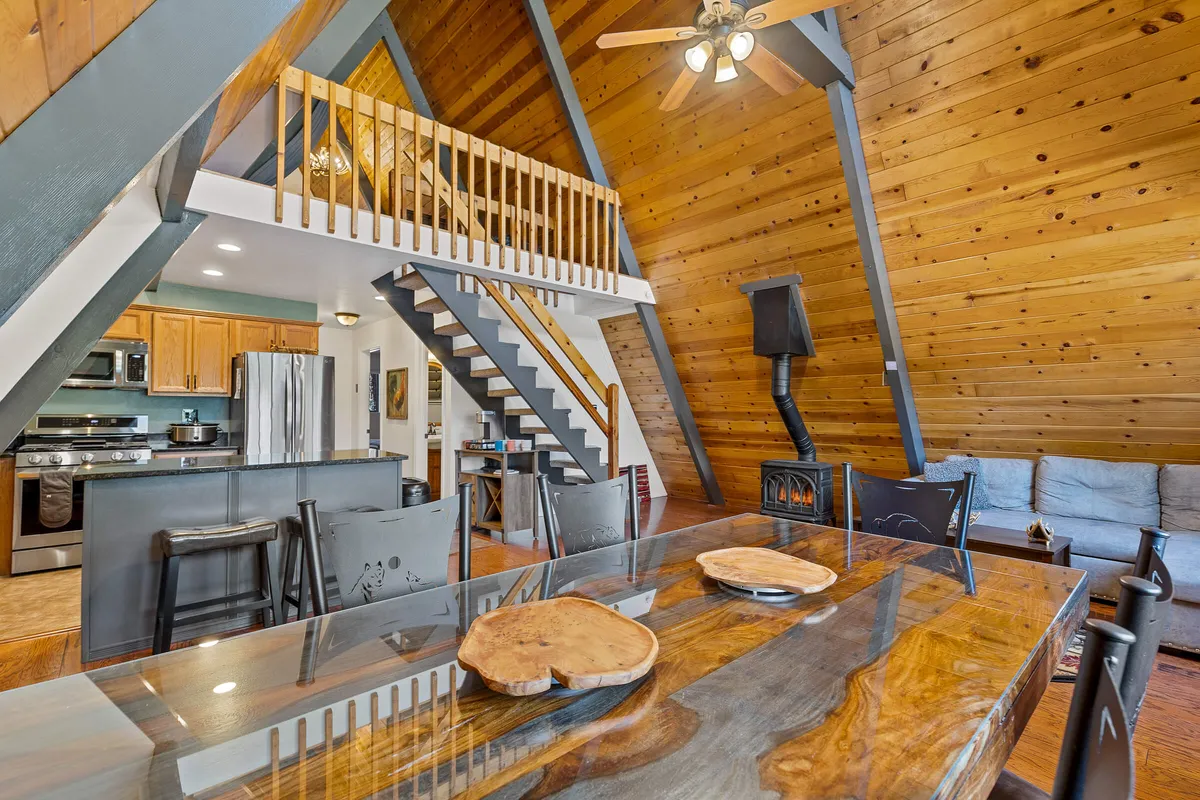 A warm and communal space showing a large wooden dining table, a staircase to a loft, a visible kitchen area with modern appliances, and a cozy wood-burning stove. The open layout is filled with natural light and designed for gathering.