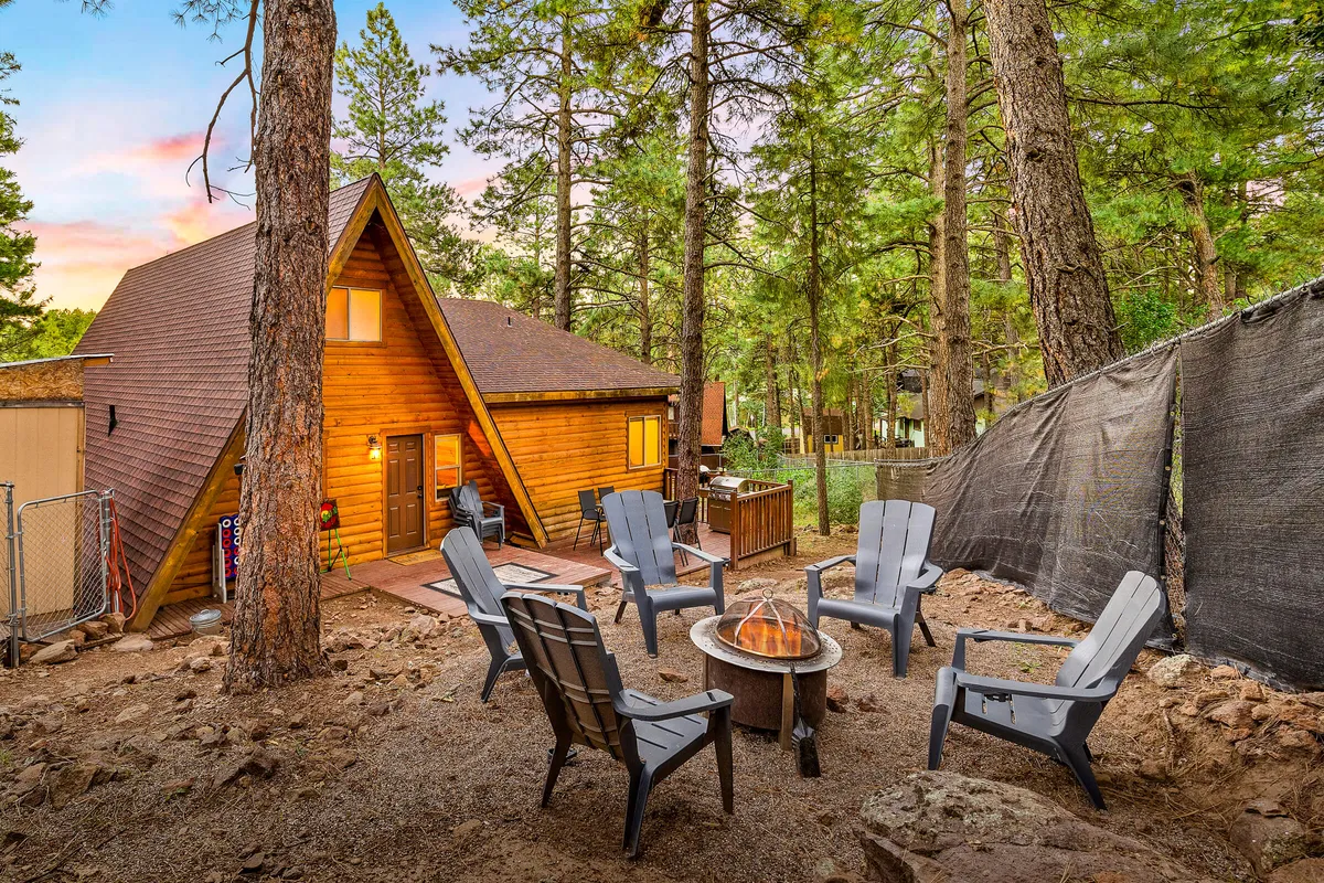 This exterior image showcases a wooden cabin surrounded by trees, with a fire pit encircled by Adirondack chairs. The setting exudes a sense of relaxed community, perfect for gathering around the fire to share stories or roast marshmallows after a day exploring the outdoors.