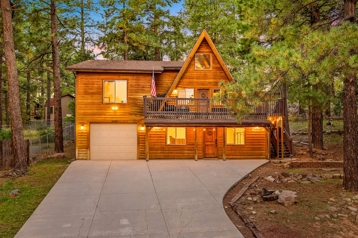 A welcoming view of the cabin's wooden exterior, complete with a spacious front deck, large windows, and an American flag. The deck is set up for outdoor gatherings, offering inviting spaces for relaxing, socializing, and enjoying the surrounding nature.