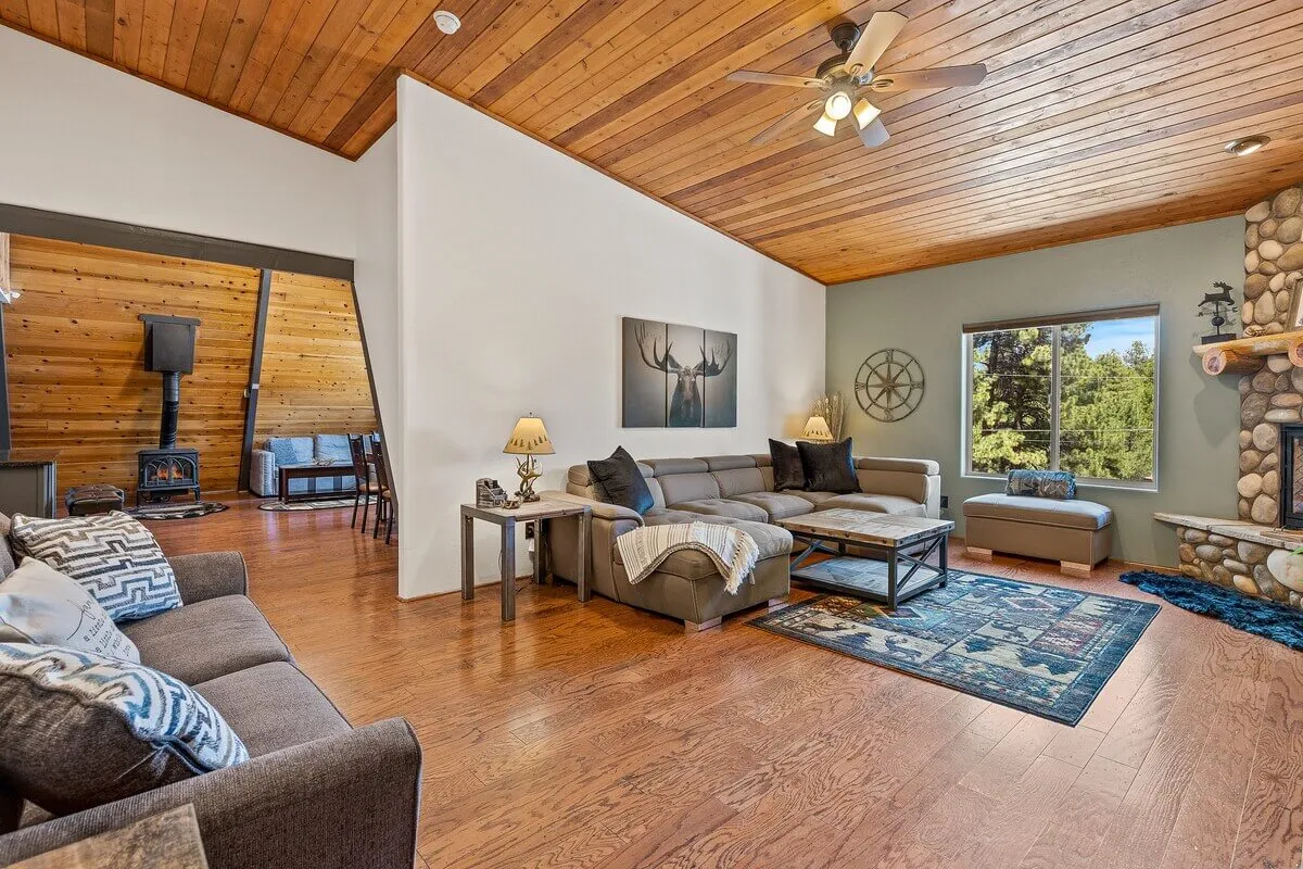 A cozy interior living space featuring a stone fireplace, comfortable seating, and a large window that lets natural light and forest views pour inside. The atmosphere is warm and welcoming, with clear sightlines connecting the interior to the natural landscape just beyond.
