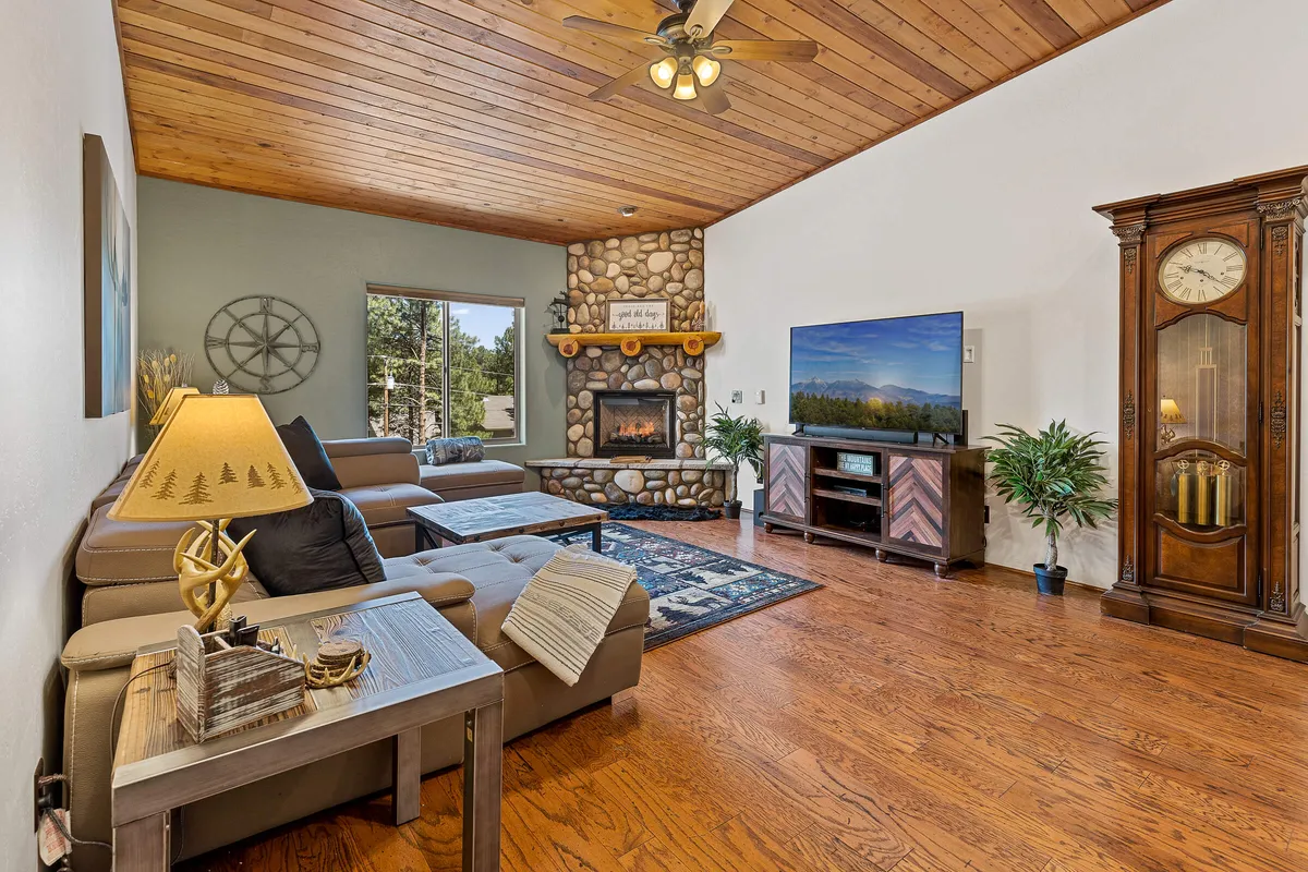 A warm, welcoming living space featuring a striking stone fireplace at the center, framed by a wooden ceiling and a large window that lets in natural light. A plush seating area surrounds the hearth, accompanied by an entertainment center and decorative touches like a clock, creating an inviting space for gatherings, relaxation, or movie nights.