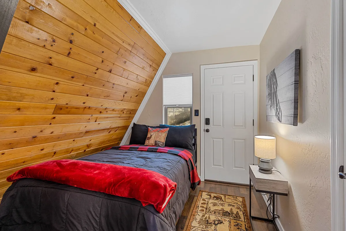 A warm, inviting bedroom featuring a wooden accent wall, a comfortable bed with a red throw blanket, artwork above the bed, and a lamp on the side table. The room exudes a balance of rustic cabin coziness and modern comfort, perfect for unwinding after a long day.